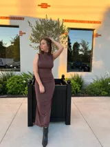 Vestido largo de color marrón, con diseño sin mangas y cuello alto. Presenta un detalle drapeado en el lateral que recorre la silueta, aportando textura y un ajuste entallado.