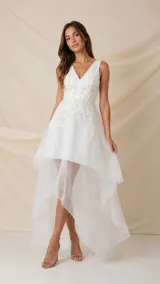 Vestido corto de tul color marfil con diseño asimétrico tipo high-low, escote en V y sin mangas. Presenta delicadas incrustaciones de flores y hojas bordadas en hilo de seda en la parte superior.