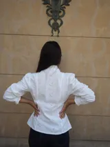 Camisa de manga larga confeccionada en broderie blanco, con diseño entallado, cuello alto y cierre frontal de botones. Presenta mangas abullonadas con hombros fruncidos y puños terminados en volados.