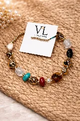 Pulsera artesanal compuesta por cuentas de piedras naturales facetadas y lisas en tonos blanco, turquesa, rojo oscuro y ámbar, intercaladas con cuentas metálicas doradas de forma irregular y cuentas pequeñas doradas. Incluye una etiqueta con el logo "VL by MARIMBA".
