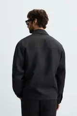 Sobrecamisa negra de corte regular, confeccionada en tejido de lino. Presenta cuello solapa, manga larga con puño abotonado y cierre frontal con cremallera metálica. Incluye bolsillos ocultos en las costuras laterales.