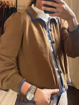 Saco de punto color marrón con detalles de costura y bordes en color azul eléctrico. Cuenta con cuello, cierre frontal con botones y mangas largas abullonadas en los hombros con puños elásticos.