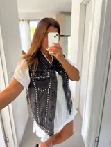 Chaleco de jean negro gastado con detalles de tachas plateadas en el frente y la espalda. Tiene cuello camisero, cierre de botones frontales y bolsillos con solapa en el pecho.