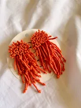 Caravanas colgantes de diseño artesanal, compuestas por una base circular densamente bordada con pequeñas cuentas y flecos largos de cuentas tubulares en color naranja vibrante.