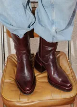 Bota estilo tejano de cuero color bordó con acabado brillante, taco cuadrado y punta cuadrada. Presenta costuras decorativas en el empeine y caña media.
