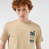 Camiseta de algodón color beige, de manga corta y cuello redondo, con diseño gráfico estampado en el pecho que incluye texto y olas estilizadas.