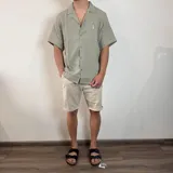 Camisa de hombre color verde oliva, de muselina con textura arrugada, manga corta, corte oversize, cuello cubano y cierre con botones. Presenta un pequeño logo bordado en el pecho.