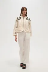 Campera estilo bomber color beige claro, con cuello redondo, cierre frontal y detalles de volados en los bordes. Presenta bordados florales en negro y blanco en la zona del pecho y bordados de tréboles negros en las mangas.