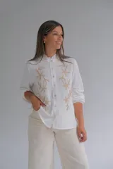 Blusa blanca de corte recto con cuello mao y volado pequeño, manga larga con puños elásticos y cierre frontal de botones. Presenta un delicado bordado floral en color beige en ambos lados del frente.