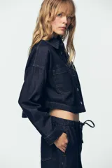 Camisa de denim color azul oscuro, con cuello solapa, manga larga y corte cropped. Presenta dos bolsillos de plastrón con tapa en el frente y cierre frontal con botones metálicos.