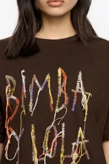 Camiseta de manga corta, corte oversize, color marrón oscuro. Presenta un estampado frontal con el texto "BIMBA Y LOLA" realizado con hilos de colores vibrantes que cuelgan, simulando un bordado texturizado.