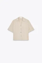 Camisa color beige de cuello solapa con escote pico y manga corta. Detalle de nido de abeja en espalda. Cierre frontal con botones.