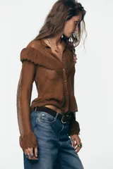 Chaqueta de punto pointelle color tostado, con cuello subido y manga larga. Presenta detalles de volantes en el pecho y puños, y cierre frontal con botones.