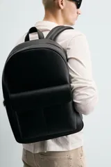 Mochila de diseño minimalista en color negro con acabado texturizado. Cuenta con un compartimento principal con cierre de cremallera, un bolsillo frontal con solapa y compartimento interior para dispositivos electrónicos. Incluye espalda acolchada, banda de sujeción para equipaje, asa de mano y correas de hombro ajustables.