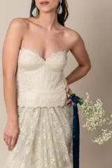 Vestido de novia strapless color natural, confeccionado en tul bordado con corsé drapeado y sobrefalda de tul. Presenta una faja con guarda en la cintura.