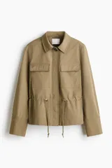 Campera en sarga de algodón color beige, con cuello, cierre frontal con botones ocultos y cordón ajustable en la cintura. Presenta bolsillos de parche con solapa en el pecho y mangas largas.