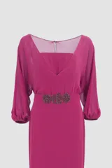 Vestido largo de fiesta color fucsia, con escote cuadrado y mangas 3/4 abullonadas. Presenta un cinturón con apliques de pedrería y una falda con paneles de gasa y volados.