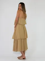 Vestido largo tipo solera de color marrón chocolate, sin mangas con tirantes finos tipo halter y detalle de aplicación de flor de tela en el escote. Presenta diseño de capas escalonadas en la falda.