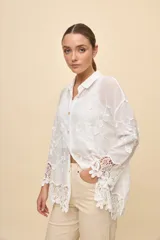Camisa blanca de calce relajado, con cuello clásico, mangas 3/4 y cierre frontal de botones. Presenta bordados florales en relieve y detalles de crochet calado en los puños y el ruedo.