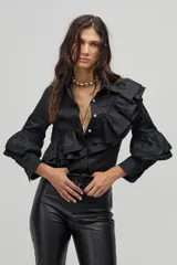Camisa negra de popelina de algodón con cuello clásico y cierre frontal de botones de nácar. Presenta volados dobles que recorren el frente y la espalda desde el hombro hasta la cintura, y volados adicionales en las mangas a la altura de los codos.