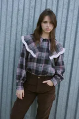 Camisa de manga larga con estampado de cuadros en tonos grises y naranjas. Presenta un cuello grande con volados negros y un corte relajado con mangas ligeramente abullonadas.