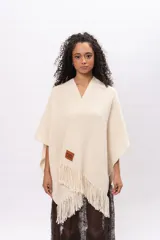 Ruana de tejido de punto en color crudo, con diseño abierto, flecos en el borde inferior y etiqueta de cuero con la marca Pura Lana en el frente.