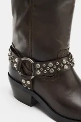 Bota de caña alta en piel color marrón oscuro, con detalle de tira con tachas y abalorios alrededor del tobillo, acabado en punta cuadrada y tacón plano.