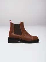 Botas tipo Chelsea de cuero color marrón, con paneles laterales elásticos en color marrón oscuro, suela de goma negra y detalle de tirador trasero en color rojo.