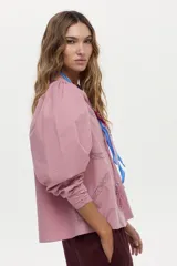 Blusa de algodón liviano color blanco, con detalles de puntilla en el frente, cuello mao con escote en V, cordón colgante y mangas largas abullonadas con puños ajustados.