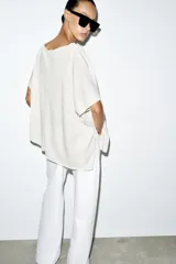 Top tipo capa blanco de punto fluido, con mezcla de viscosa, cuello bote, mangas cortas y bajo asimétrico.
