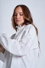 Camisa de color azul con cuello alto decorado con volados y lazo de ajuste. Presenta tablas decorativas en los hombros, cierre frontal con botones de nácar y mangas largas con puños elásticos que generan un efecto abullonado.