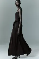 Vestido largo de color negro con escote recto y tirantes finos ajustables. Presenta un diseño con detalle de volados en la cintura que genera un efecto peplum y una falda con caída fluida.