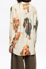 Camisa de corte recto con cuello clásico, cierre frontal de botones y manga larga con puños abotonados. Presenta un estampado integral de perros salchicha sobre fondo color crema.