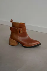 Bota corta de cuero negro con acabado combinado mate y charol. Presenta una tira decorativa con tachas metálicas y hebilla en el tobillo, tacón cuadrado robusto y suela con plataforma.