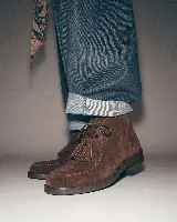 Bota de caña media confeccionada en cuero gamuzado color marrón chocolate. Presenta un diseño de estilo mocasín con cordones frontales decorados con borlas desmontables y suela plana de goma resistente.