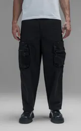 Pantalón cargo oversize de corte relajado, confeccionado en textil técnico resistente de color negro. Presenta bolsillos laterales cargo de gran tamaño con cierres metálicos, bolsillos laterales superiores y bolsillos traseros con broches a presión. Incluye cinturón integrado de cinta de seguridad con hebilla de encastre técnica.