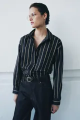 Camisa de corte oversize confeccionada en mezcla de algodón, con diseño de rayas verticales en tonos azul marino y beige. Presenta cuello solapa, cierre frontal oculto con botones, manga larga con puños y un bolsillo en el pecho. El bajo es asimétrico.