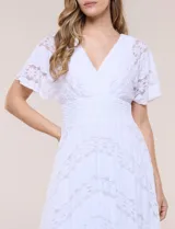 Vestido midi blanco de estilo romántico confeccionado en poliéster con elastano, con detalles de encaje floral. Cuenta con escote cruzado en V, mangas cortas acampanadas, cintura elastizada y falda evasé con terminación asimétrica en picos.