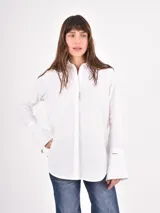 Camisa blanca de corte clásico con cuello camisero y puños anchos destacados. Presenta cierre frontal oculto por tapeta y dobladillo redondeado.