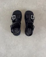 Sandalias de cuero color marrón con suela de goma negra, tiras anchas y hebilla metálica.