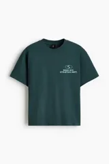 Remera boxy de punto liviano de algodón en color verde oscuro, de hombros caídos, con cuello redondo acanalado y bajo recto. Presenta un diseño estampado en el pecho con texto y logotipo.
