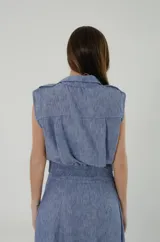 Top de lino mezcla con poliéster, color celeste melange, de corte chaleco con cuello camisero, sin mangas y abotonado al frente con botones de pasta efecto nácar. Cuenta con pretina lisa en la delantera, elástico ancho en la espalda, canesú con pliegues y charreteras en los hombros.