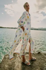 Vestido largo estilo túnica de color crudo con estampado multicolor de motivos marinos y mediterráneos. Presenta cuello con volante, cierre frontal con botones, manga larga y cordón ajustable en el cuello con abalorios.