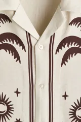 Camisa de manga corta color beige, con cuello abierto y cierre frontal de botones. Presenta un estampado en marrón de palmeras, soles y estrellas, con líneas en los bordes.
