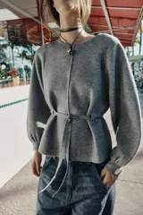 Jersey de punto color gris, con cuello redondo, mangas largas abullonadas con puños elásticos y cintura ajustable mediante cordón. Presenta una costura vertical marcada en la parte frontal.