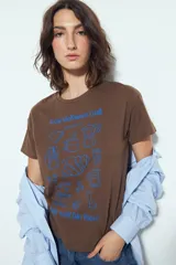 Remera marrón de manga corta con estampado frontal en azul que incluye la frase "Slow Mornings Club" y dibujos relacionados con el café y el desayuno, como una cafetera, tazas, granos de café y un croissant.