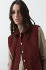 Chaleco de pana color marrón oscuro, con cuello camisero, cierre frontal de botones metálicos dorados y dos bolsillos frontales con detalle de bordado geométrico.