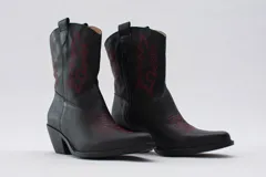 Botas estilo cowboy de cuero negro, caña media, con taco ancho y punta ligeramente afilada. Presentan bordados decorativos en hilo rojo en la caña y en la puntera.