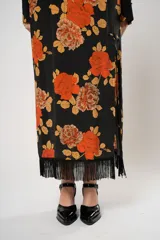 Vestido largo negro con estampado de flores naranjas y amarillas, cuello mao con abertura en forma de gota, mangas largas acampanadas con botones en los puños, tajo lateral y flecos en el ruedo.