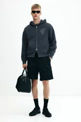 Campera con capucha de algodón pesado, color azul oscuro o gris pizarra, con cierre frontal bidireccional y bolsillos canguro. Presenta un corte holgado (boxy) con hombros caídos y puños y bajo acanalados.